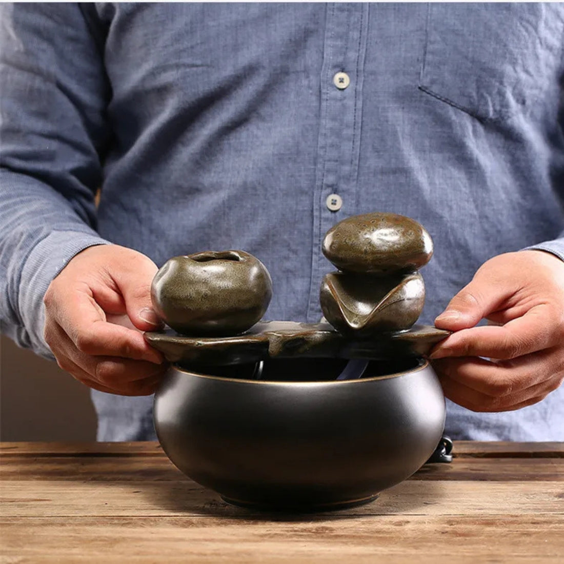 Personne tenant les composants d'une fontaine zen artisanale montrant sa taille compacte, avec un bassin circulaire en céramique noire et des éléments d'écoulement texturés.