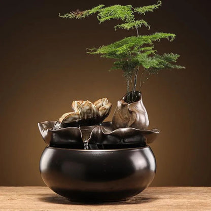 Fontaine zen d'intérieur en céramique avec écoulement d'eau sur une fleur de lotus sculptée, pot intégré avec plante verte délicate et base noire mate.
