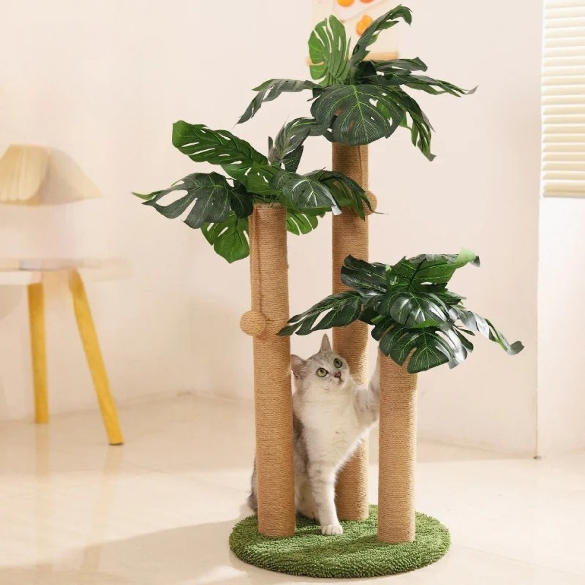 Arbre à chat design en forme de plante Monstera avec trois poteaux griffoirs en corde de chanvre, feuilles tropicales artificielles et base circulaire verte, avec un chat gris jouant à l'intérieur.