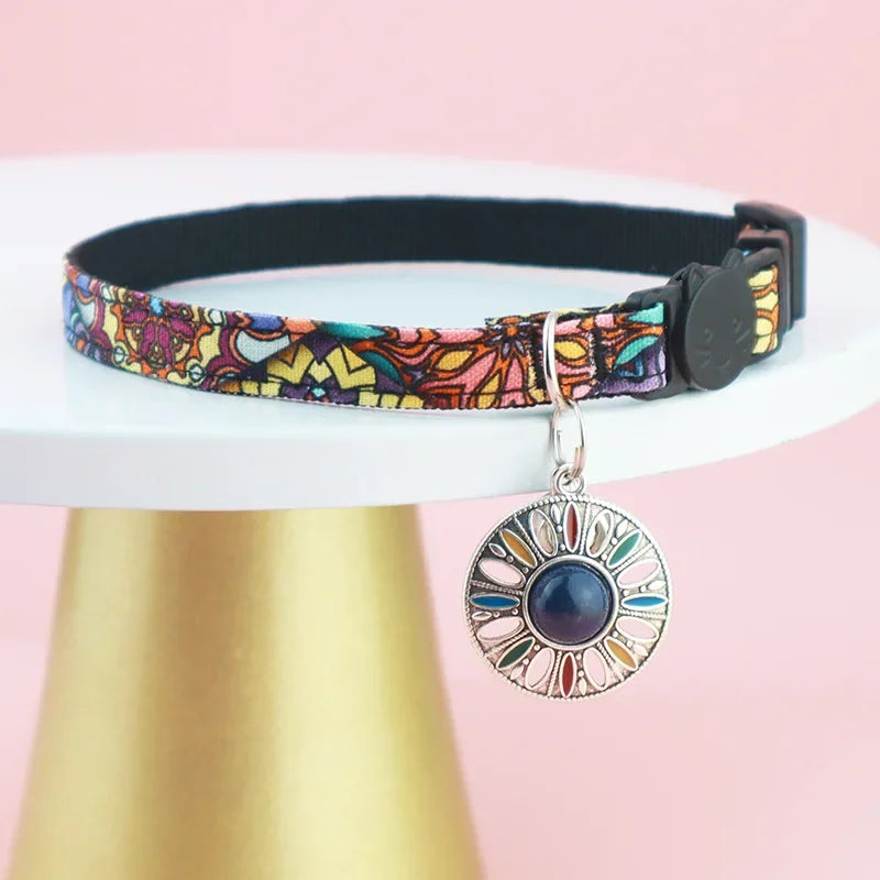 Bijou de cou pour chat au motif floral violet et jaune, présenté sur un support doré élégant, avec un médaillon mandala orné d'une pierre centrale bleu foncé et un fermoir de sécurité noir.