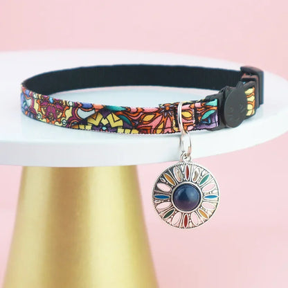 Bijou de cou pour chat au motif floral violet et jaune, présenté sur un support doré élégant, avec un médaillon mandala orné d'une pierre centrale bleu foncé et un fermoir de sécurité noir.