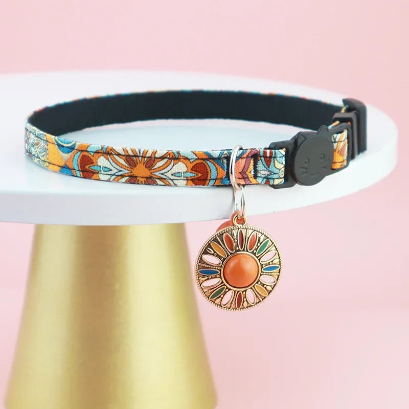 Collier pour chat de luxe au motif floral orange et turquoise, présenté sur un support doré élégant, avec un grand pendentif mandala soleil et un fermoir noir tête de chat.