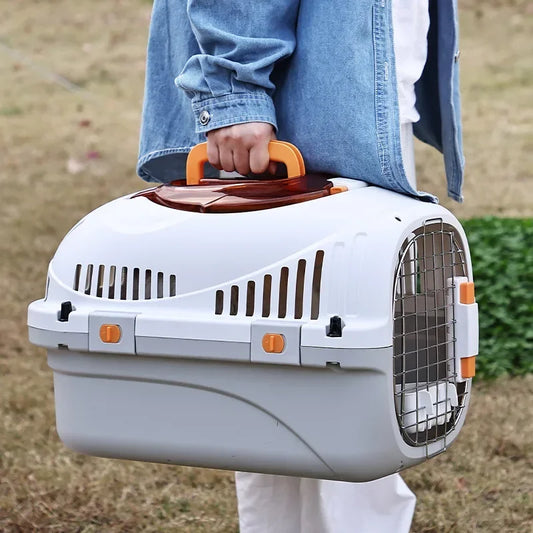 Caisse de transport rigide pour chat gris et blanc, dotée d'une grille de sécurité métallique, de verrous latéraux orange robustes et d'une poignée de transport ergonomique, portée à la main en extérieur.