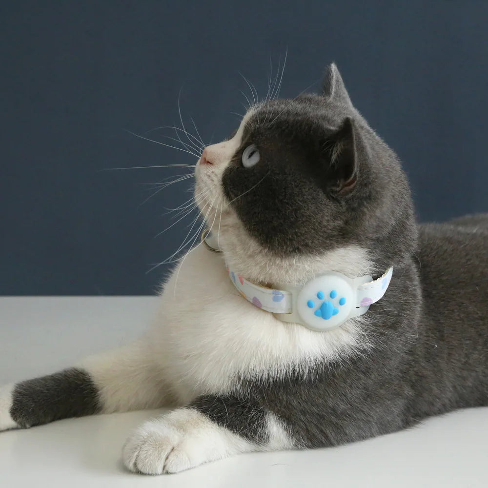 Chat bicolore gris et blanc allongé portant un collier gps pour chat blanc à motifs cœurs pastels. Gros plan sur le support AirTag en silicone blanc avec empreinte de patte bleue, parfaitement positionné. Ce collier gps chat esthétique et léger offre une protection sécurisée. Un accessoire type collier gps à chat Kawaii et confortable pour la protection quotidienne de votre félin.
