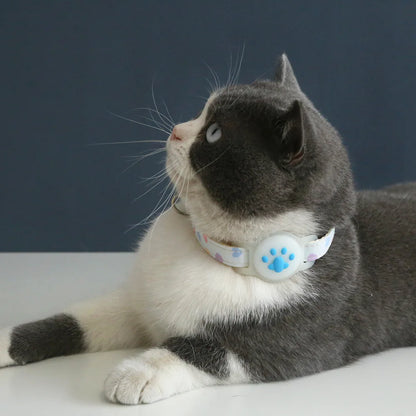 Chat bicolore gris et blanc allongé portant un collier gps pour chat blanc à motifs cœurs pastels. Gros plan sur le support AirTag en silicone blanc avec empreinte de patte bleue, parfaitement positionné. Ce collier gps chat esthétique et léger offre une protection sécurisée. Un accessoire type collier gps à chat Kawaii et confortable pour la protection quotidienne de votre félin.