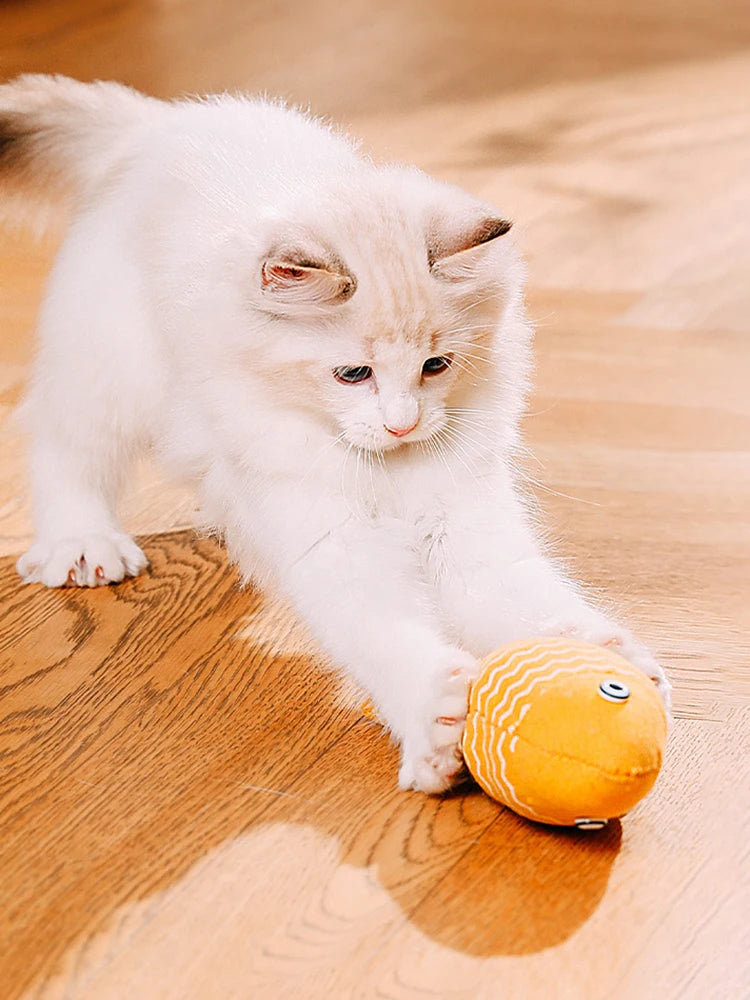 Chaton blanc et beige en pleine action de jeu, attrapant avec ses griffes un poisson en tissu jaune à motifs zigzag blancs sur un parquet en bois.