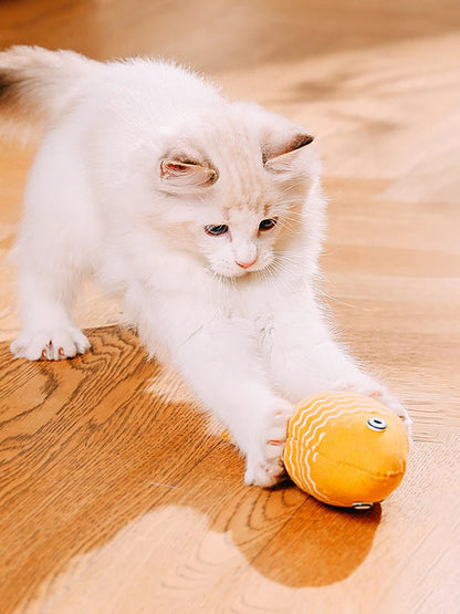 Chaton blanc et beige en pleine action de jeu, attrapant avec ses griffes un poisson en tissu jaune à motifs zigzag blancs sur un parquet en bois.
