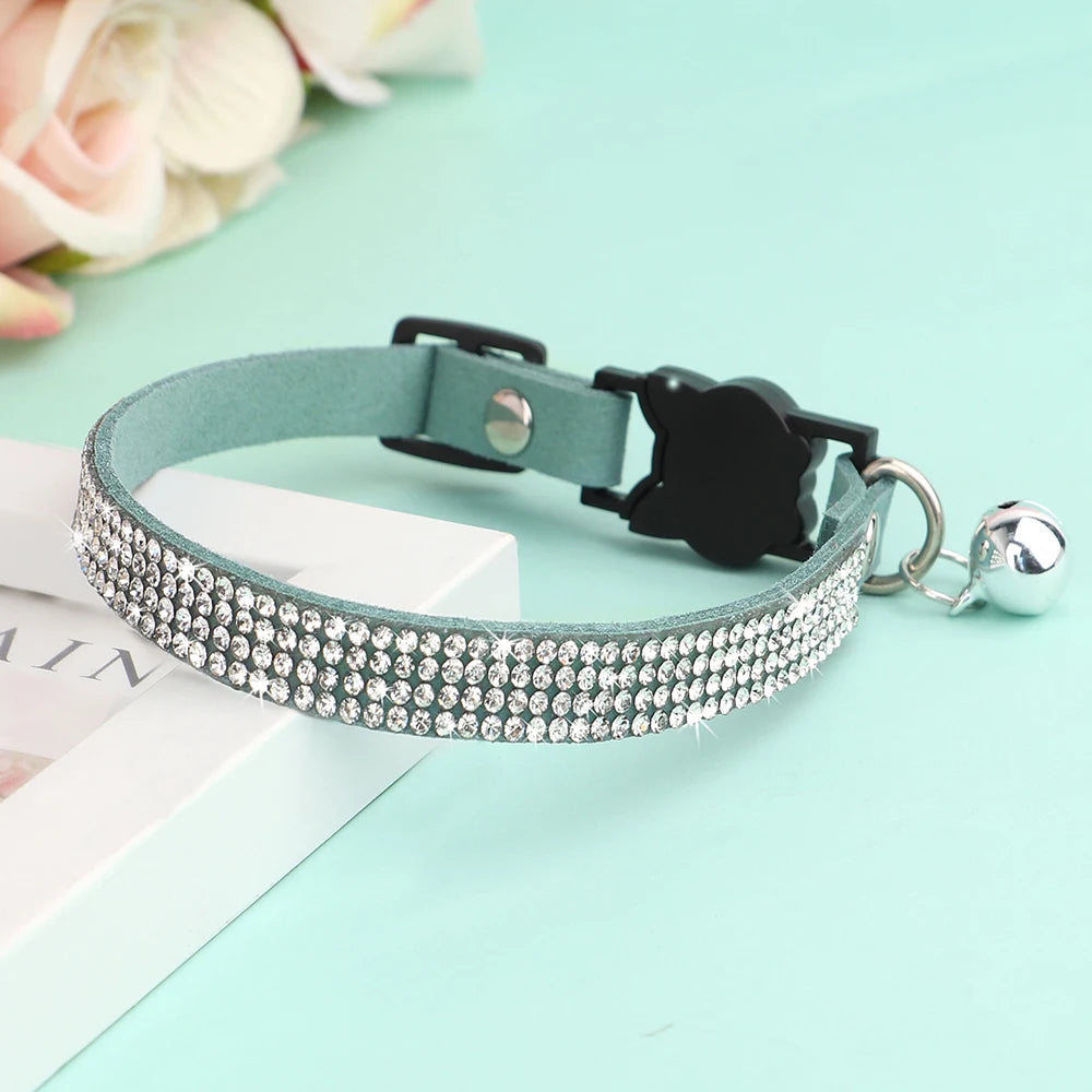 Collier pour chat en velours bleu menthe orné de trois rangées de strass étincelants, avec boucle de sécurité noire et grelot argenté posé sur un support élégant.