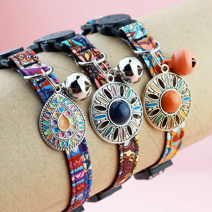 Zoom sur trois colliers pour chats aux motifs ethniques colorés, présentés sur un support cylindrique simulant le cou, avec médaillons mandala détaillés et clochettes argentées et cuivrées.