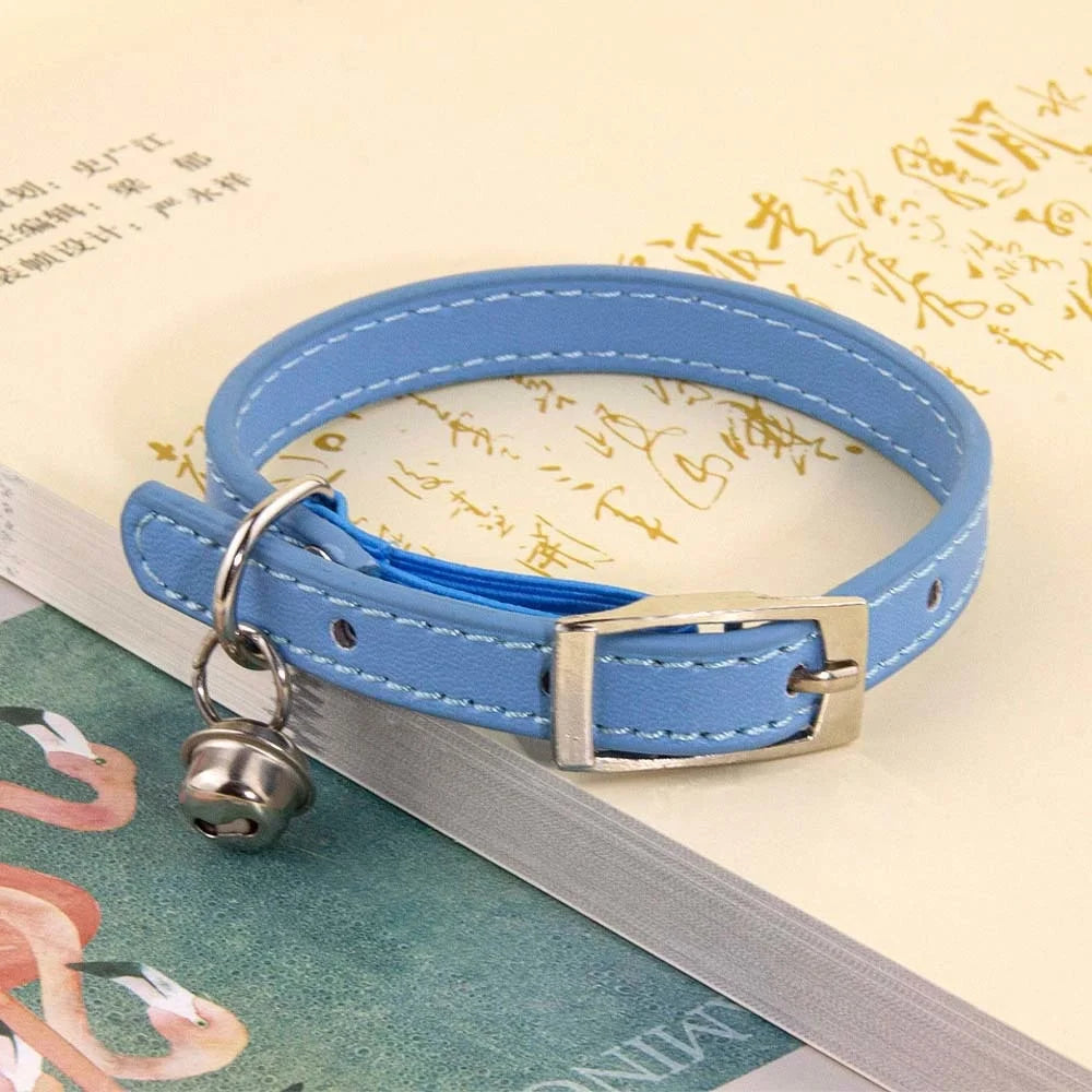 Collier pour chat en cuir bleu ciel avec surpiqûres blanches, doté d'une boucle ardillon en métal argenté et d'un petit grelot, posé sur un livre ouvert.