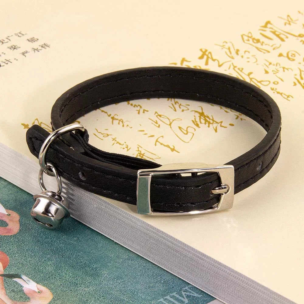 Collier pour chat en cuir noir avec surpiqûres ton sur ton, doté d'une boucle ardillon en métal argenté et d'un grelot assorti, posé sur un livre ancien ouvert.