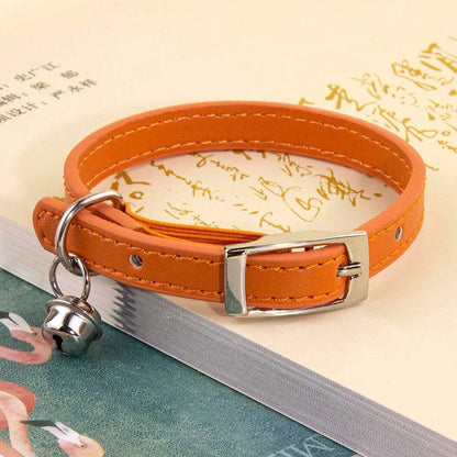 Collier pour chat en cuir orange mandarine avec surpiqûres ton sur ton, doté d'une boucle ardillon en métal argenté et d'un petit grelot, posé sur un livre ouvert.