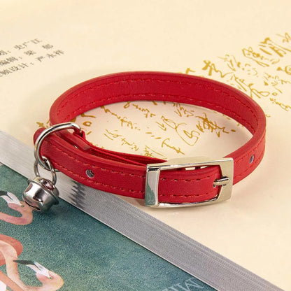 Collier pour chat en cuir rouge avec surpiqûres ton sur ton, doté d'une boucle ardillon en métal argenté et d'un petit grelot, posé sur un livre ouvert.