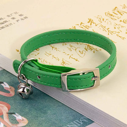 Collier pour chat en cuir vert émeraude avec surpiqûres ton sur ton, doté d'une boucle ardillon en métal argenté et d'un petit grelot, posé sur un livre ancien ouvert.