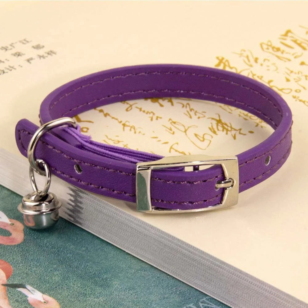 Collier pour chat en cuir violet avec surpiqûres ton sur ton, doté d'une boucle ardillon en métal argenté et d'un grelot assorti, posé sur un livre ancien ouvert.
