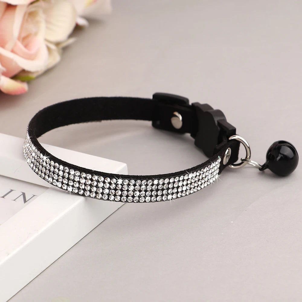 Collier pour chat en velours noir orné de trois rangées de strass étincelants, doté d'une boucle de sécurité noire en forme de tête de chat et d'un grelot noir assorti posé sur un support blanc.