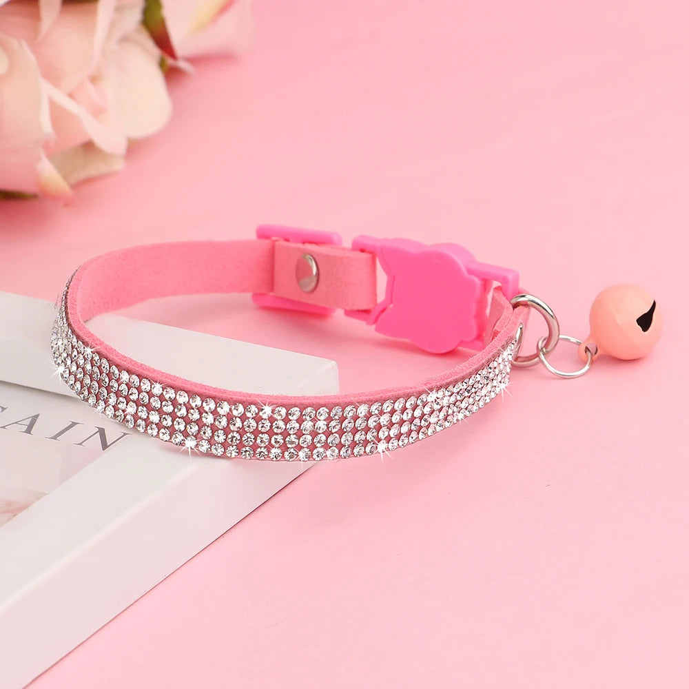 Collier pour chat en velours rose orné de trois rangées de strass étincelants, avec boucle de sécurité rose et grelot assorti posé sur un support élégant.