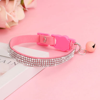 Collier pour chat en velours rose orné de trois rangées de strass étincelants, avec boucle de sécurité rose et grelot assorti posé sur un support élégant.