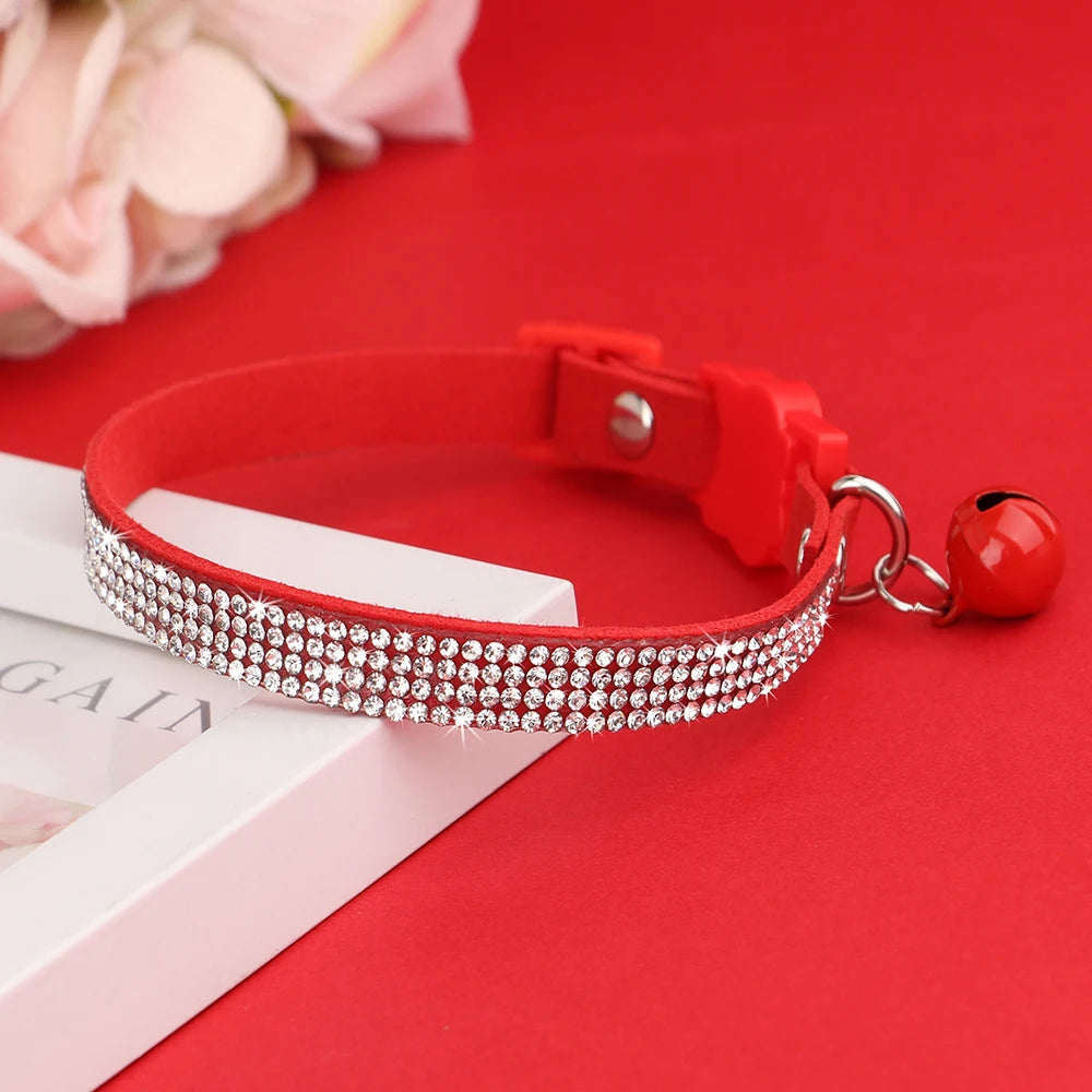 Collier pour chat en velours rouge vif orné de trois rangées de strass étincelants, avec boucle de sécurité et grelot rouge assorti posé sur un support élégant.