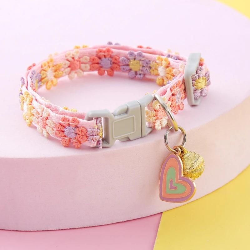 Collier pour chat rose aux fleurs brodées dans les tons pastel (lilas, abricot). Ce collier chaton ajustable dispose d'un pendentif cœur multicolore et d'une clochette dorée. Accessoire de style bohème-chic pour collier chat, alliant design artisanal et sécurité. Finitions premium Les Trésors d'Owka.