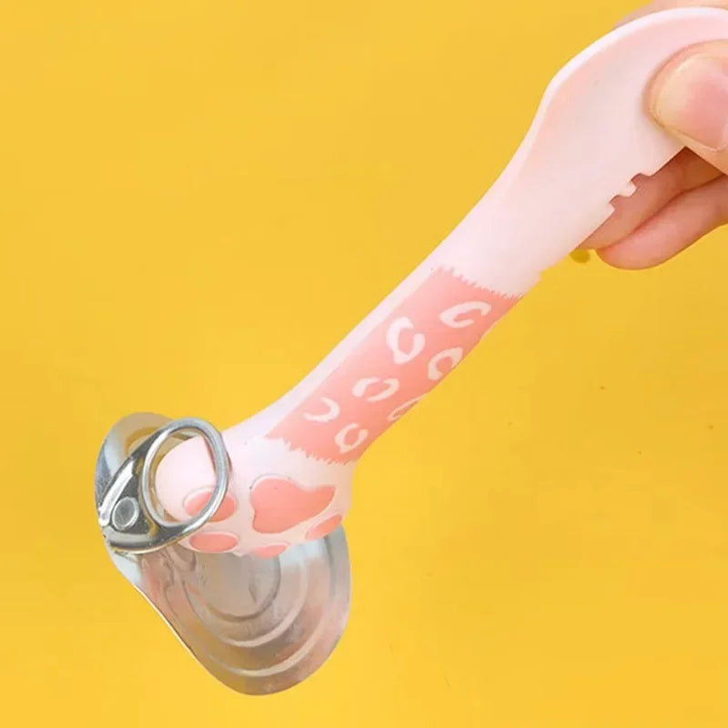 Main utilisant une cuillère ergonomique Lucky Cat rose pour ouvrir facilement l'opercule d'une boîte de pâtée pour chat, démonstration de la fonction levier sur fond jaune.