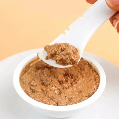Gros plan d'une cuillère à pâtée blanche pour chat servant une portion de nourriture humide, montrant les bords crantés ergonomiques pour un raclage parfait des boîtes de conserve.