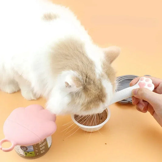 Chat blanc et roux mangeant de la pâtée servie avec une cuillère ergonomique blanche à motif patte rose, accompagnée d'un couvercle hermétique en silicone en forme de patte pour boîte de conserve.