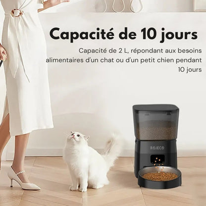 Distributeur automatique ROJECO noir de 2 litres offrant une autonomie de 10 jours pour chat ou petit chien, montrant un chat blanc attendant son repas à côté de l'appareil.