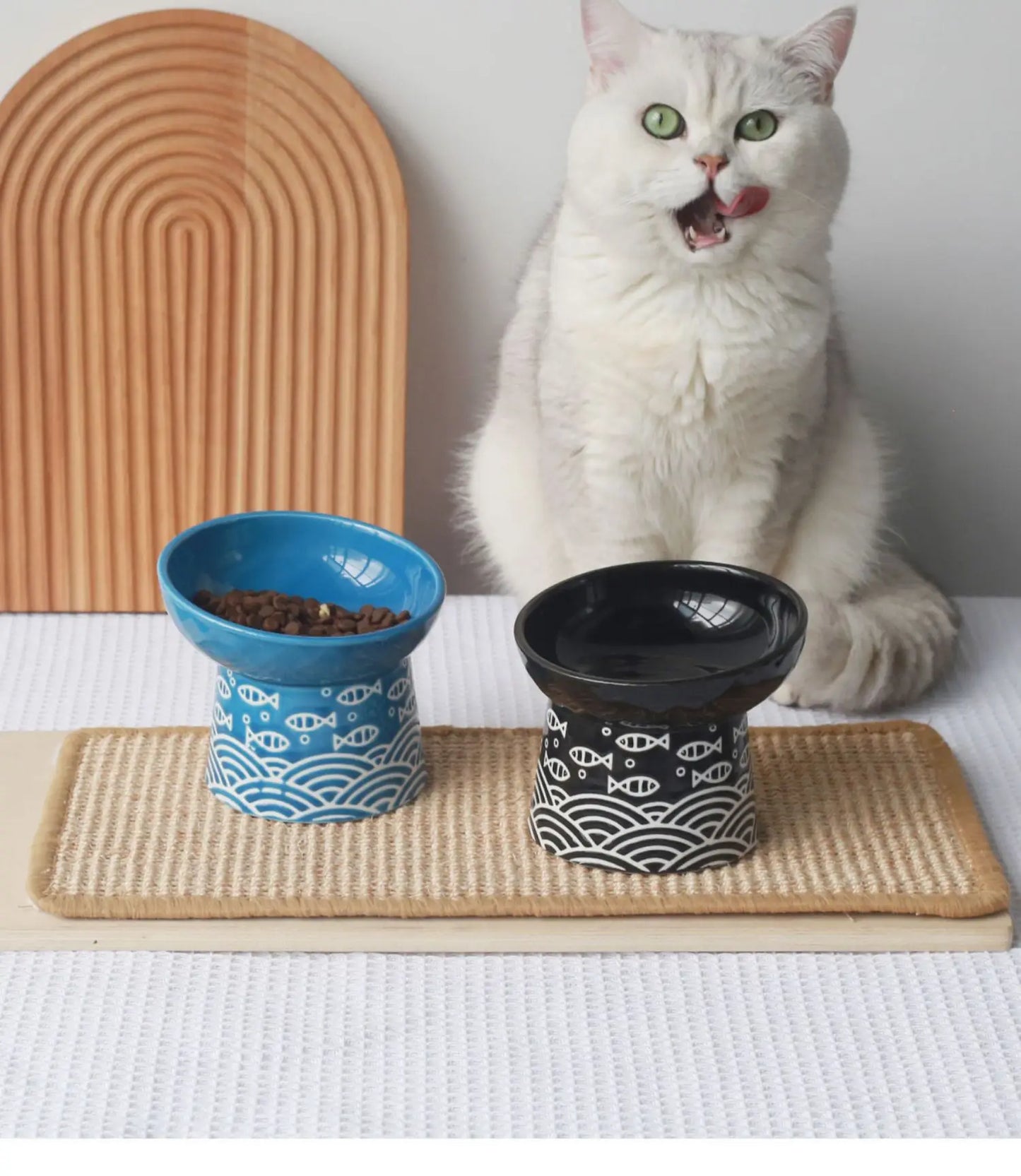 Chat blanc dégustant ses croquettes dans un ensemble de gamelle pour chat surélevée en céramique bleue et noire au design japonais motif poisson. Cette gamelle chat orthopédique de luxe sur socle haut assure une posture ergonomique et une hygiène parfaite. Une gamelle stable et élégante posée sur un tapis naturel pour le bien-être de votre animal.