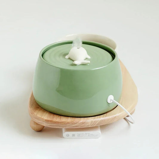 Fontaine à eau pour chat en céramique vert sauge de forme ronde, avec une buse centrale en forme de petite baleine blanche diffusant un jet d'eau doux, posée sur un socle en bois naturel.