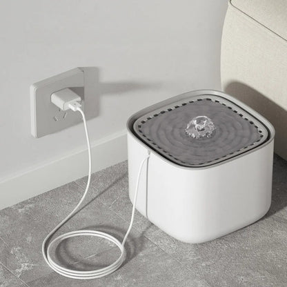Fontaine à eau pour chat carrée blanche branchée sur une prise secteur murale, avec jet d'eau central bouillonnant et câble USB blanc discret.