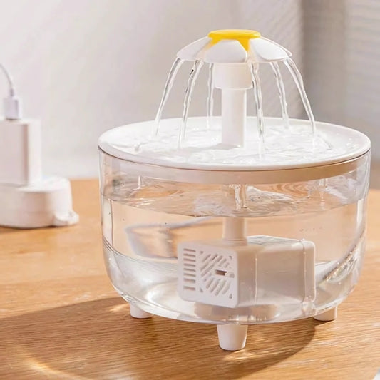 Fontaine à eau pour chat entièrement transparente avec buse marguerite, montrant la pompe immergée et le niveau d'eau cristalline, posée sur une table en bois ensoleillée.