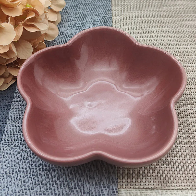 Gamelle pour chat en céramique rose poudré en forme de fleur avec bords festonnés, design peu profond pour le confort des moustaches, posée sur un tapis de table bicolore.