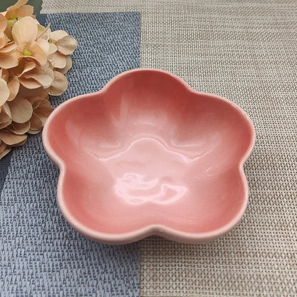 Gamelle pour chat en céramique rose corail brillant en forme de fleur, design évasé et peu profond pour prévenir la fatigue des moustaches, présentée sur un tapis de table texturé.