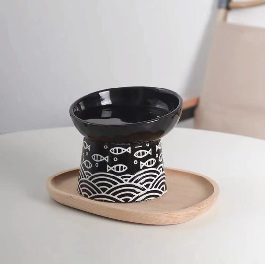 Gamelle pour chat surélevée en céramique noire avec motifs de poissons et vagues japonaises, posée sur un plateau en bois. Cette gamelle chat orthopédique design assure une posture ergonomique et une hygiène parfaite grâce à sa conception en céramique lourde et stable. Une gamelle de luxe anti-vomi pour un intérieur zen et élégant.