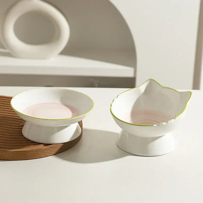 Duo de gamelle pour chat surélevée en céramique blanche avec liseré vert et fond rose pastel. Cette gamelle chat design en forme de tête de félin offre une position ergonomique pour préserver les cervicales. Une gamelle de luxe en céramique lourde et stable, parfaite pour une décoration d'intérieur moderne et minimaliste.