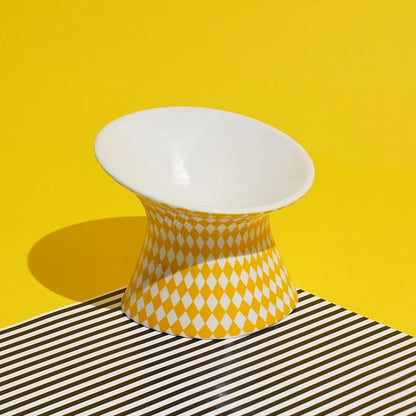 Gamelle pour chat surélevée en céramique jaune et blanche à motif losanges arlequin. Cette gamelle animaux ergonomique et inclinée à 15° protège les cervicales et facilite la digestion de votre félin. Une gamelle pour chat de luxe, stable, hygiénique et design, idéale pour un intérieur moderne et le bien-être animal.