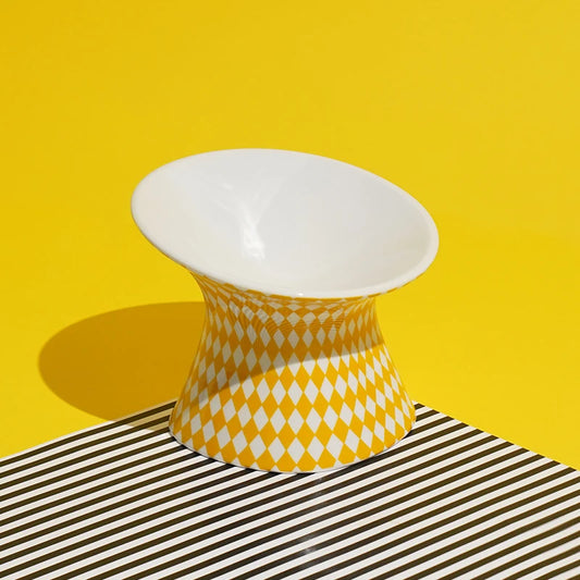 Gamelle pour chat surélevée en céramique jaune et blanche à motif losanges arlequin. Cette gamelle animaux ergonomique et inclinée à 15° protège les cervicales et facilite la digestion de votre félin. Une gamelle pour chat de luxe, stable, hygiénique et design, idéale pour un intérieur moderne et le bien-être animal.