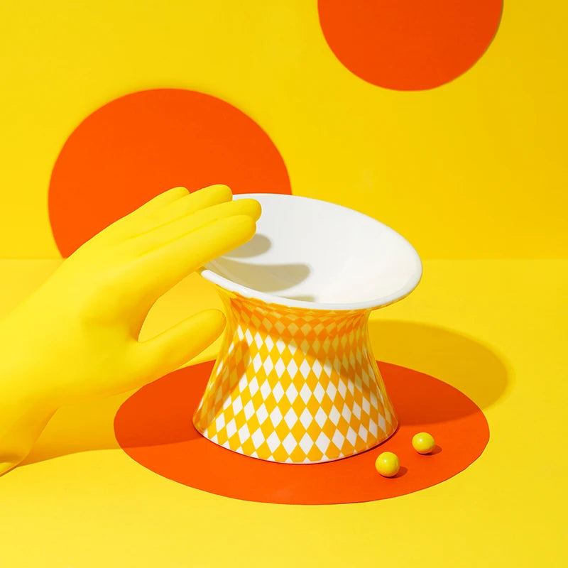 Gamelle pour chat surélevée en céramique jaune et blanche avec motif losanges arlequin. Cette gamelle animaux ergonomique et inclinée prévient les douleurs au cou et facilite la digestion de votre félin. Une gamelle pour chat au design pop-art, hygiénique et stable, idéale pour un intérieur moderne et coloré.