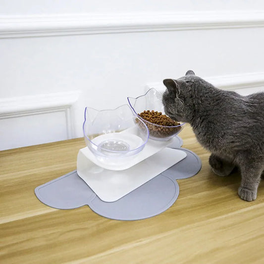 Chat gris mangeant dans une gamelle pour chat double inclinée à 15 degrés avec bols transparents design oreilles de chat. Cette gamelle chat orthopédique repose sur un tapis gris en forme de nuage pour une hygiène parfaite et un confort cervical optimal. Une gamelle élégante et ergonomique pour préserver la santé de votre animal.