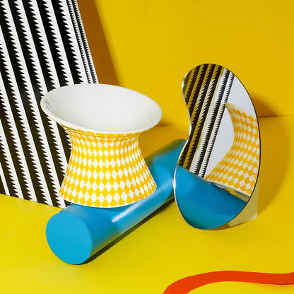 Gamelle pour chat surélevée en céramique jaune et blanche avec motif losanges arlequin dans un décor design pop. Cette gamelle animaux ergonomique et inclinée protège le cou et facilite la digestion de votre félin. Une gamelle pour chat de luxe, hygiénique, stable et artistique, idéale pour sublimer votre intérieur tout en prenant soin de votre animal.