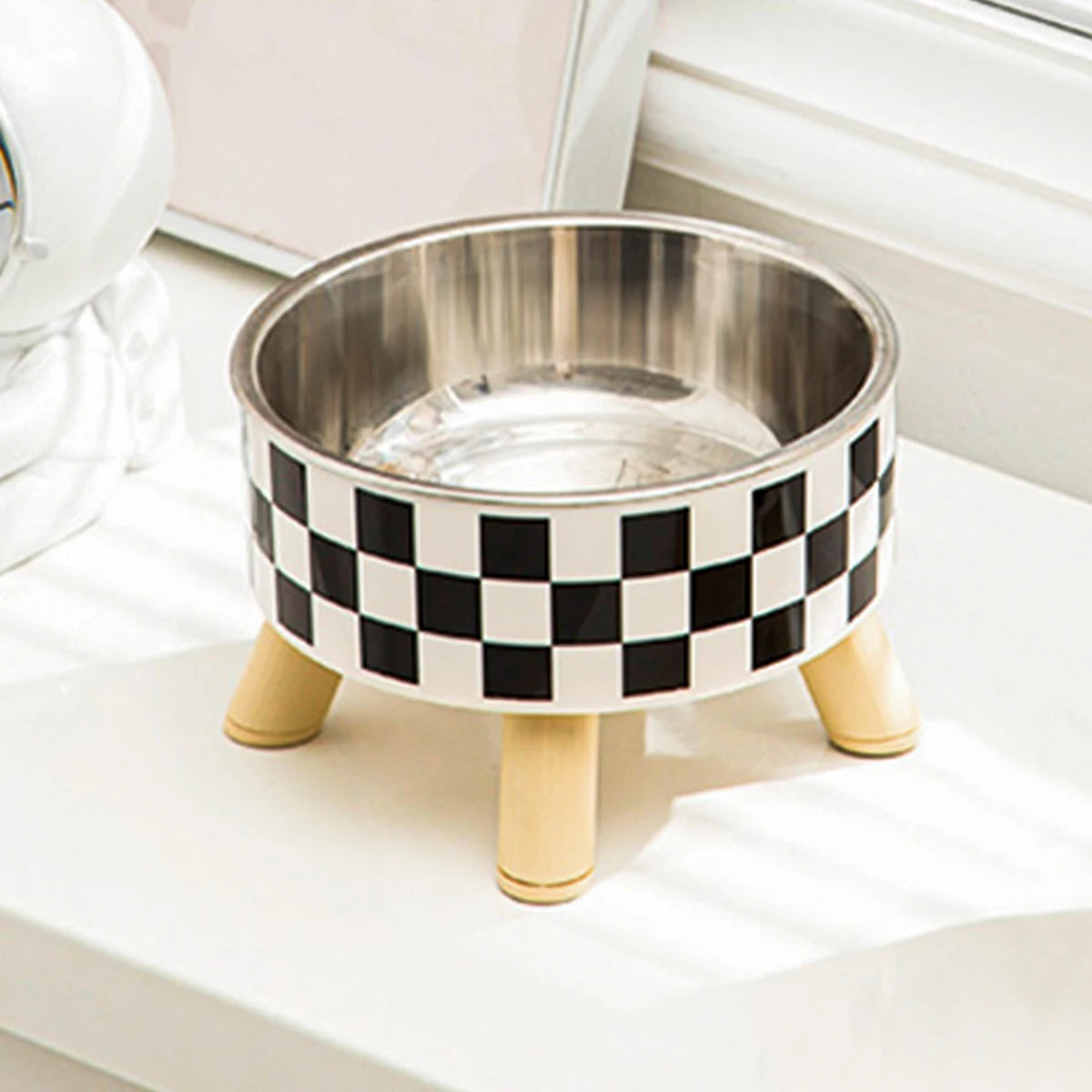 Gamelle pour chat surélevée avec bol en acier inoxydable et support trépied en bois clair au design damier noir et blanc. Cette gamelle chat orthopédique de luxe assure un confort cervical optimal et une hygiène parfaite contre l'acné du menton. Une gamelle robuste et élégante au style scandinave pour le bien-être de votre félin.
