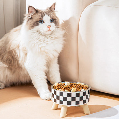 Chat Ragdoll à côté d'une gamelle pour chat surélevée avec bol en acier inoxydable et support trépied en bois clair au design damier noir et blanc. Cette gamelle chat orthopédique de luxe assure un confort cervical optimal et une hygiène parfaite contre l'acné du menton. Une gamelle robuste et élégante remplie de nourriture pour le bien-être de votre félin.