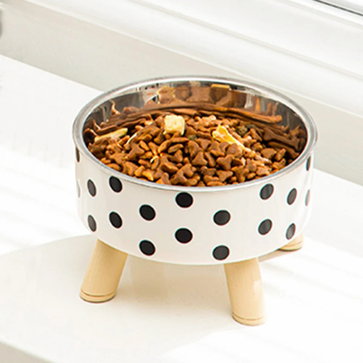 Gamelle pour chat surélevée sur pieds en bois clair avec bol en acier inoxydable et design blanc à pois noirs. Cette gamelle chat orthopédique allie style scandinave et confort digestif pour votre animal. Une gamelle robuste et élégante remplie de croquettes, idéale pour une décoration d'intérieur moderne.