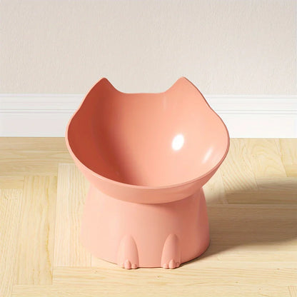 Gamelle surélevée pour chat au design ergonomique couleur corail, bol incliné en forme de tête de chat avec oreilles, posé sur un parquet clair devant un mur blanc.
