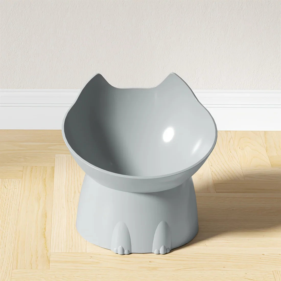 Gamelle surélevée pour chat au design ergonomique gris clair, bol incliné en forme de tête de chat avec oreilles, posé sur un parquet clair devant un mur blanc.
