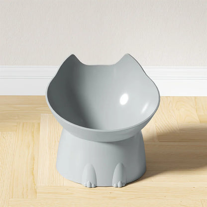 Gamelle surélevée pour chat au design ergonomique gris clair, bol incliné en forme de tête de chat avec oreilles, posé sur un parquet clair devant un mur blanc.