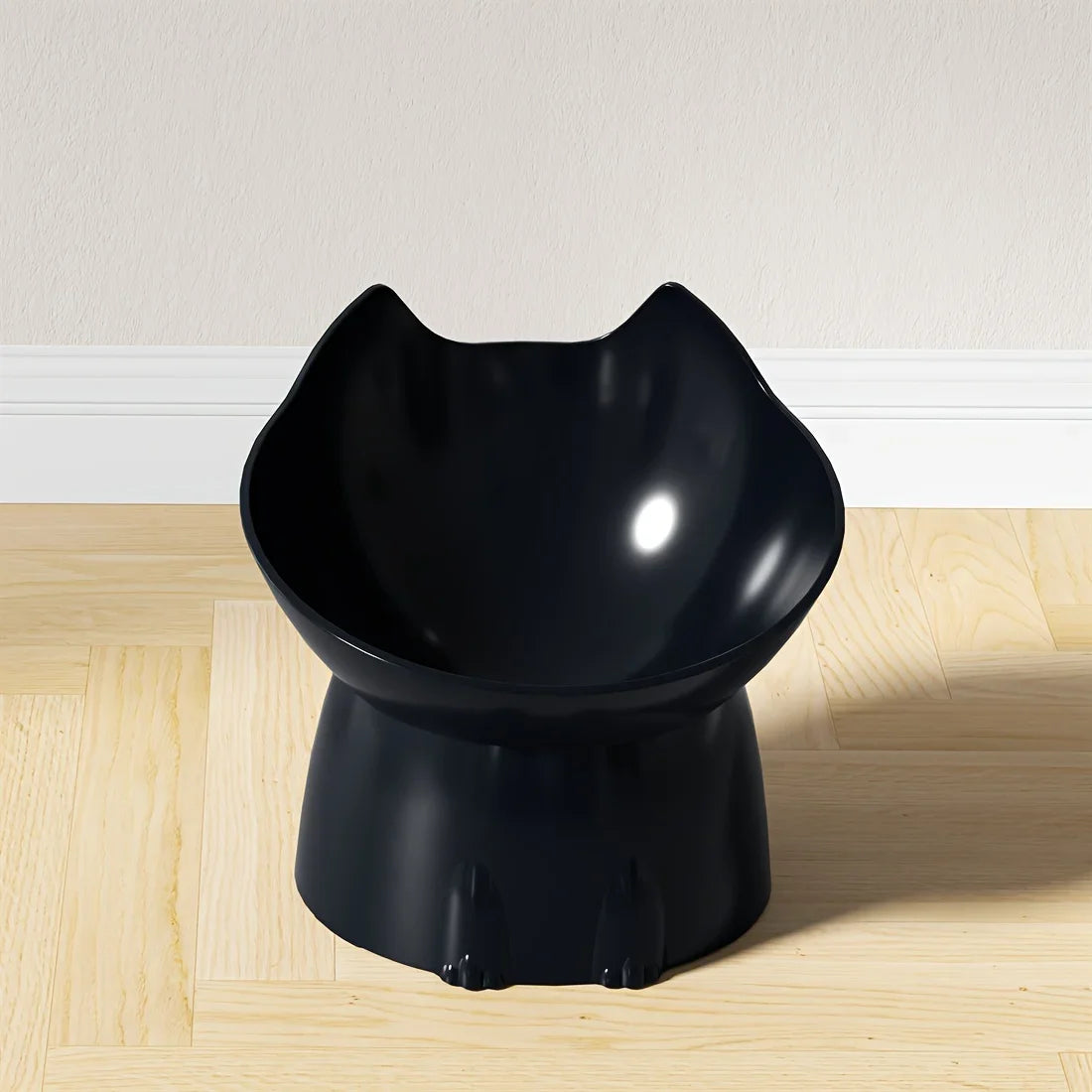 Gamelle surélevée pour chat au design ergonomique noir brillant, bol incliné en forme de tête de chat avec oreilles, posé sur un parquet clair devant un mur blanc.