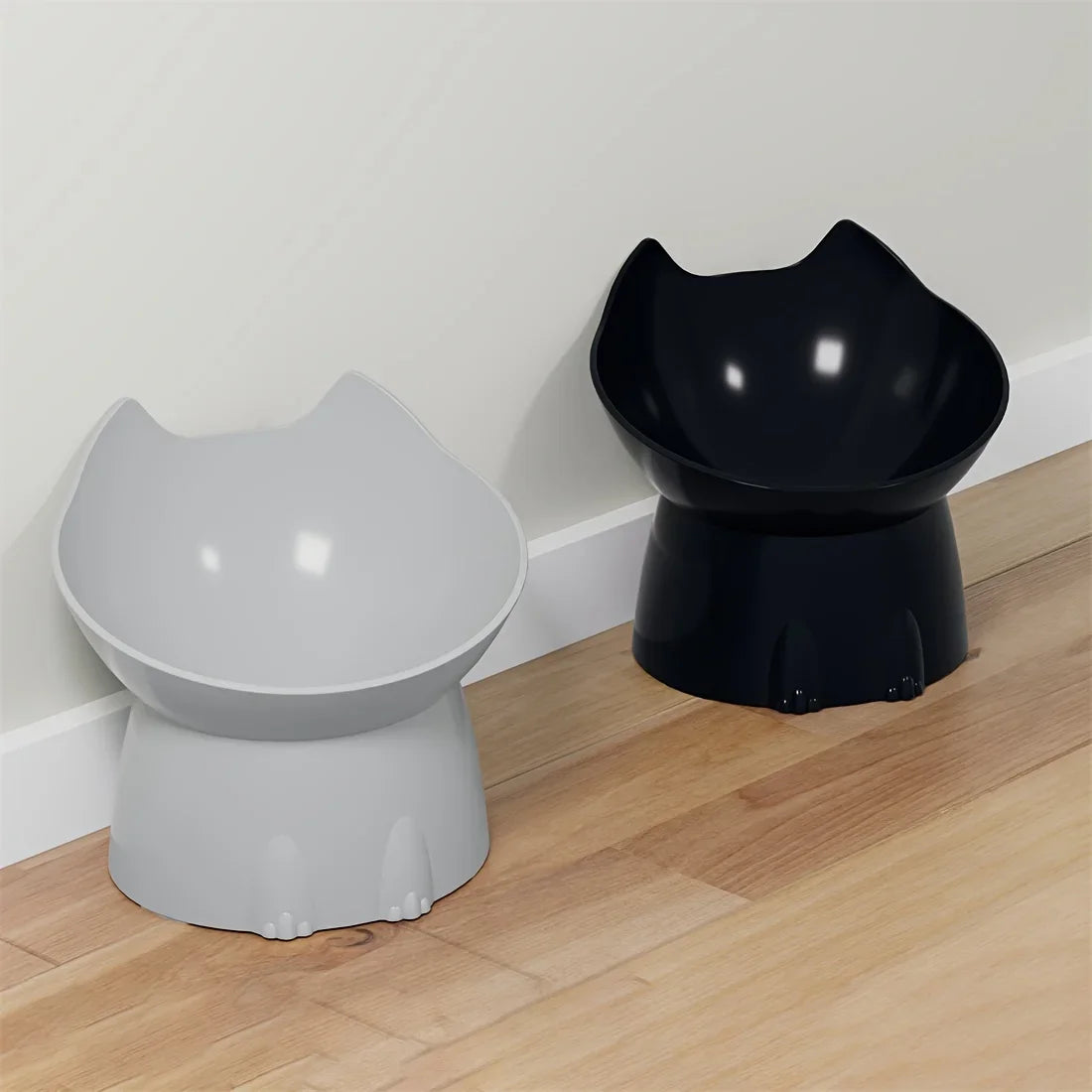 Duo de gamelles surélevées pour chat au design ergonomique avec oreilles, présentées en coloris noir brillant et gris perle le long d'un mur blanc dans un intérieur moderne