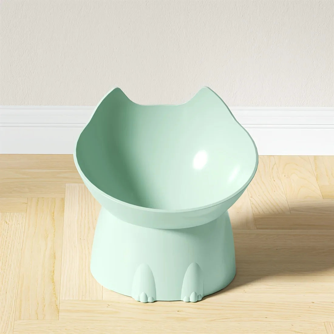 Gamelle surélevée pour chat au design ergonomique vert menthe pastel, bol incliné en forme de tête de chat avec oreilles, posé sur un parquet clair devant un mur blanc.